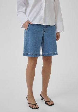 Persona che indossa pantaloncini di denim blu di lunghezza media, camicia bianca abbottonata e sandali neri con tacco, in piedi con una mano in tasca.