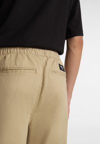 Béžové kalhoty s elastickým pasem, které mají dvě zadní kapsy a štítek s logem. Hladká tkanina, uvolněný střih a minimální designové detaily.