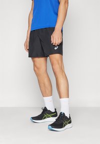 Pantalones cortos deportivos negros con un logo lateral, combinados con zapatillas de correr negras con acentos verdes y una suela blanca, y calcetas blancas.