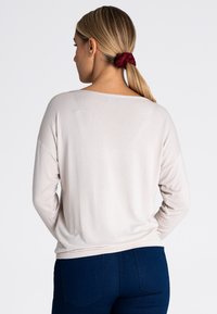 Beżowa bluzka z długim rękawem, z szerokim dekoltem, luźny krój i ribbowany dół. Połączona z ciemnoniebieskimi dżinsami. Włosy związane w kok za pomocą czerwonej gumki.