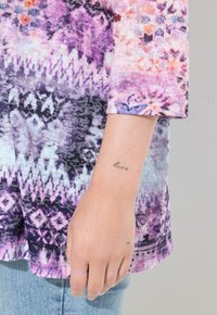 Bunt gemustertes Oberteil in lila, pink und blau. Mit einem strukturierten Design und einem sichtbaren Tattoo am Handgelenk. Kurzarm.
