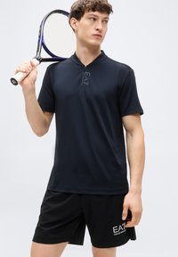 Πολο μπλούζα ναυτικού μπλε με ημικάμινο φερμουάρ και λογότυπο, σε συνδυασμό με μαύρο σορτς με λεπτομέρειες λογότυπου, κατάλληλα για αθλητικές ή καθημερινές εμφανίσεις.