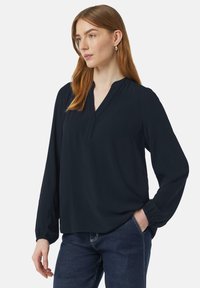 Granatowa bluzka z długim rękawem, w dekolcie w kształcie litery V i luźnym kroju. Wykonana z miękkiej tkaniny, z elastycznymi mankietami i płaskimi szwami. Noszona z ciemnymi dżinsami.