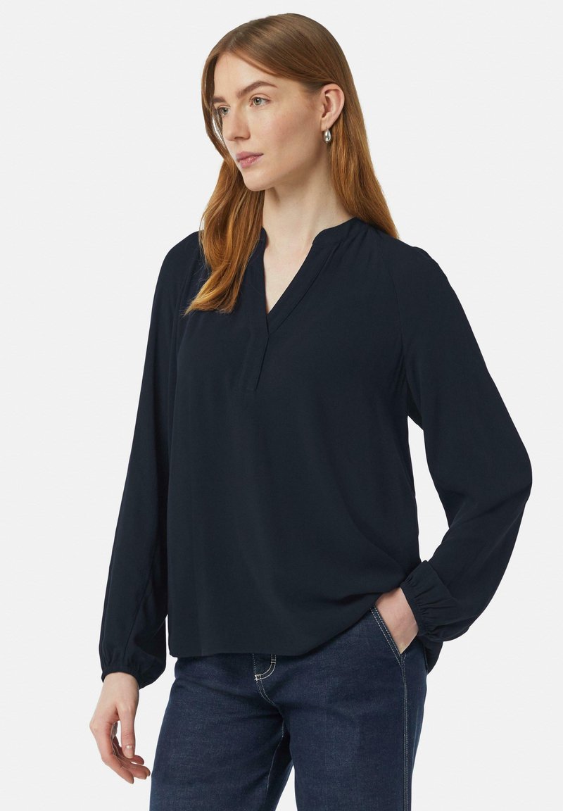 Granatowa bluzka z długim rękawem, w dekolcie w kształcie litery V i luźnym kroju. Wykonana z miękkiej tkaniny, z elastycznymi mankietami i płaskimi szwami. Noszona z ciemnymi dżinsami.