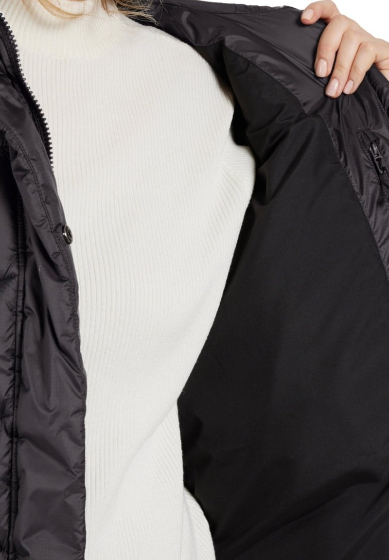 Chaqueta de plumas negra con un exterior brillante, con un cuello alto y cierre de cremallera, sobre un cuello de tortuga blanco de canalé. Tejido texturizado visible.