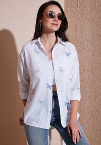 White button-up shirt with blue and pink butterfly embroidery, rolled sleeves, and a relaxed fit, paired with denim jeans.