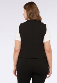 Gilet nero senza maniche dal design aderente, con dettagli delle cuciture posteriori e texture liscia, indossato sopra una camicia bianca. Aspetto semplice e ben rifinito.