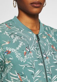 Veste bomber brodée en sarcelle avec motifs floraux et oiseaux. Comprend une fermeture éclair à l'avant et un col côtelé, mettant en valeur un tissu texturé.