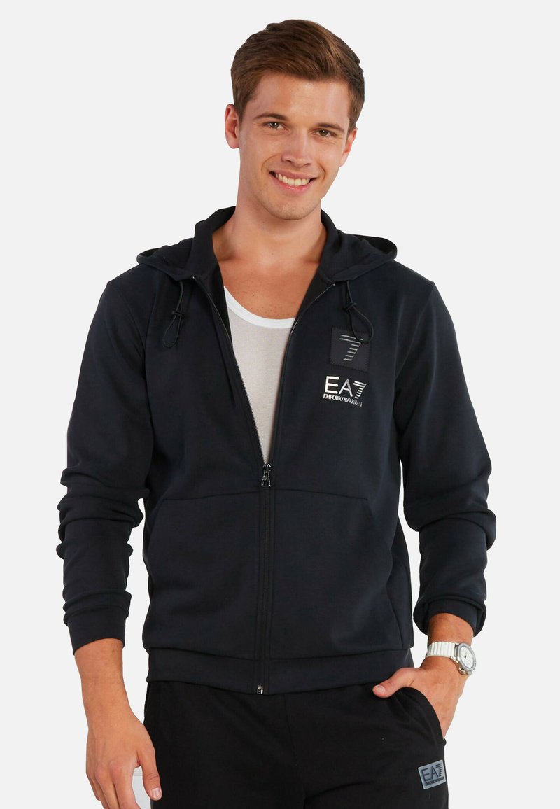 Felpa nera con zip realizzata in tessuto liscio, dotata di cappuccio con coulisse, tasche frontali e un piccolo logo sul petto.
