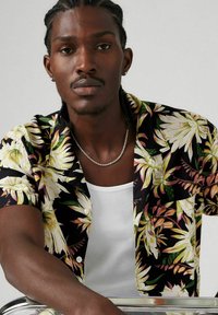 Camisa de manga corta con estampado floral en negro, con flores amarillas y rosas, que presenta un collar y botones. Lleva encima una camiseta sin mangas de canalé blanca.