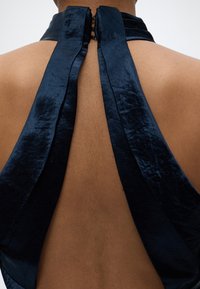 Vue de dos d'une personne portant un vêtement noir en velours avec un col licou, le haut du dos découvert et une fermeture à agrafe.