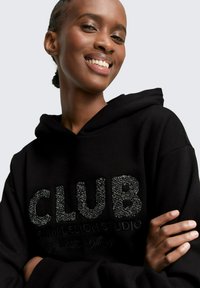 Schwarzer Hoodie mit "CLUB" in strukturierten, glitzernden Buchstaben, mit einer Kordelzugkapuze und weichem Stoff. Das Modell zeigt ein Lächeln und hat die Arme verschränkt.