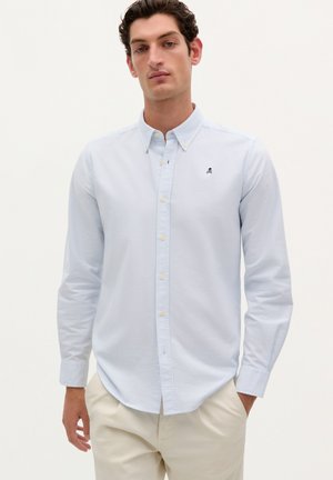 Camicia azzurra chiara con righe verticali, maniche lunghe e un piccolo logo ricamato sul petto. Design casual e sartoriale.