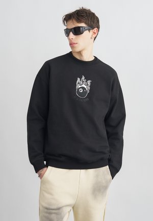 Νεαρός άνδρας φοράει μαύρα γυαλιά ηλίου, μαύρο φούτερ με γραφικό φλεγόμενο μπάλα και μπεζ παντελόνι με τα χέρια στις τσέπες.