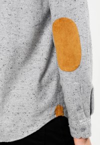 Šedý svetr s černými skvrnami, s hnědými semišovými náplety na loktech a knoflíkovým manžetou na rukávech. Odolná tkanina s uvolněným střihem.