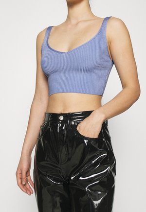 Persona con top corto de canalé lavanda y pantalones de vinilo negro brillante, con una mano en el bolsillo, sobre un fondo liso.