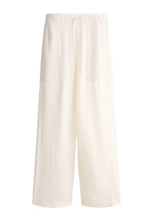 Kobieta ubrana w luźną, kremową odzież domową i czarne sandały, niosąca dużą paskowaną torbę, chodząca po sześciokątnej podłodze z płytek.