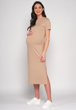 Zwangere vrouw in beige midi-jurk en witte sneakers, zijwaarts staand met één hand op haar buik, glimlachend tegen een effen achtergrond.