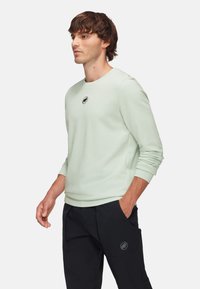 Mint green long-sleeve sweatshirt with a round neckline and a small black logo on the chest. Smooth texture, paired with dark pants.