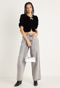 Giacca cropped in velluto nero con maniche a sbuffo e dettaglio a fiocco, abbinata a jeans grigi a vita alta e a gamba larga, e una borsa argentata.