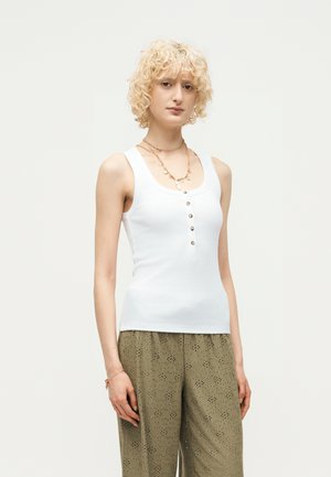 Personne blonde aux cheveux bouclés portant un haut blanc sans manches boutonné et un pantalon large vert olive à motif œillet.