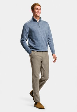 Lächelnder Mann geht, trägt blauen Pullover mit Viertel-Reißverschluss, beige Hose und braune Chelsea-Stiefel aus Wildleder vor schlichtem weißem Hintergrund.