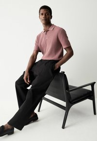 Lichtroze geribd poloshirt met kraag en drie knopen, gecombineerd met losse zwarte broek en zwarte instapschoenen, zittend op een zwarte stoel.