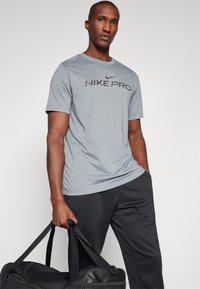 Grå Nike Pro t-skjorte med rund hals, korte ermer og en svart logo; kombinert med mørke bukser og en svart treningspose.