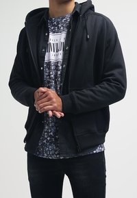 Černá mikina na zip s kapucí a stahovací šňůrkou, pod ní vzorované šedé tričko. Má žebrované manžety a pohodlný střih. Vytvořeno s tmavými džínami.