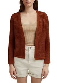 Rust-colored knitted cardigan with open front, long sleeves, and a textured pattern, worn over a beige ribbed tank top and white shorts.
