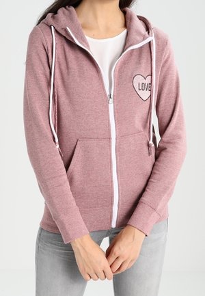 Femme portant un sweat à capuche rose zippé avec une fermeture éclair blanche, des cordons pour la capuche, des poches devant et un cœur avec le texte "LOVE" sur la poitrine.