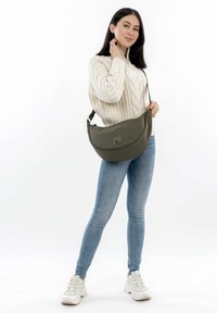 Borsa verde oliva con forma curva e texture liscia, caratterizzata da un piccolo dettaglio logo. Il modello indossa un maglione crema e jeans chiari.