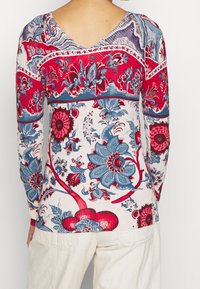 T-shirt à manches longues avec un col en V, arborant un motif floral rouge et bleu sur un fond clair, fabriqué en tissu doux et texturé.
