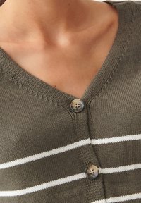 Zbliżenie na brązowy dziergany sweter z białymi, poziomymi paskami oraz dwoma marmurkowymi guzikami w okolicy obojczyka.