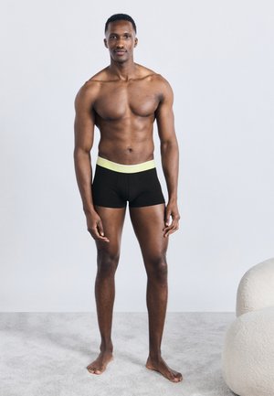 Μυώδης άνδρας όρθιος ξυπόλητος πάνω σε χαλί, φοράει μαύρα εσώρουχα μποξέρ με ανοιχτό κίτρινο λουρί μέσης, ουδέτερο φόντο, χαλαρή στάση.