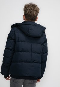 Dunkelblaue Steppjacke mit Kapuze, ausgestattet mit einer gesteppte Textur und elastischen Bündchen. Auf dem Rücken befindet sich ein Logo-Patch auf der linken Schulter.