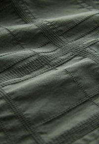 Grüner Stoff mit einem strukturierten Gittermuster, der dezente Nähte und eine glatte, matte Oberfläche aufweist.