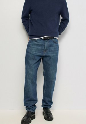 Hombre con vaqueros azules, cinturón negro, zapatos de cuero negros y suéter azul marino de pie contra una pared lisa y clara.