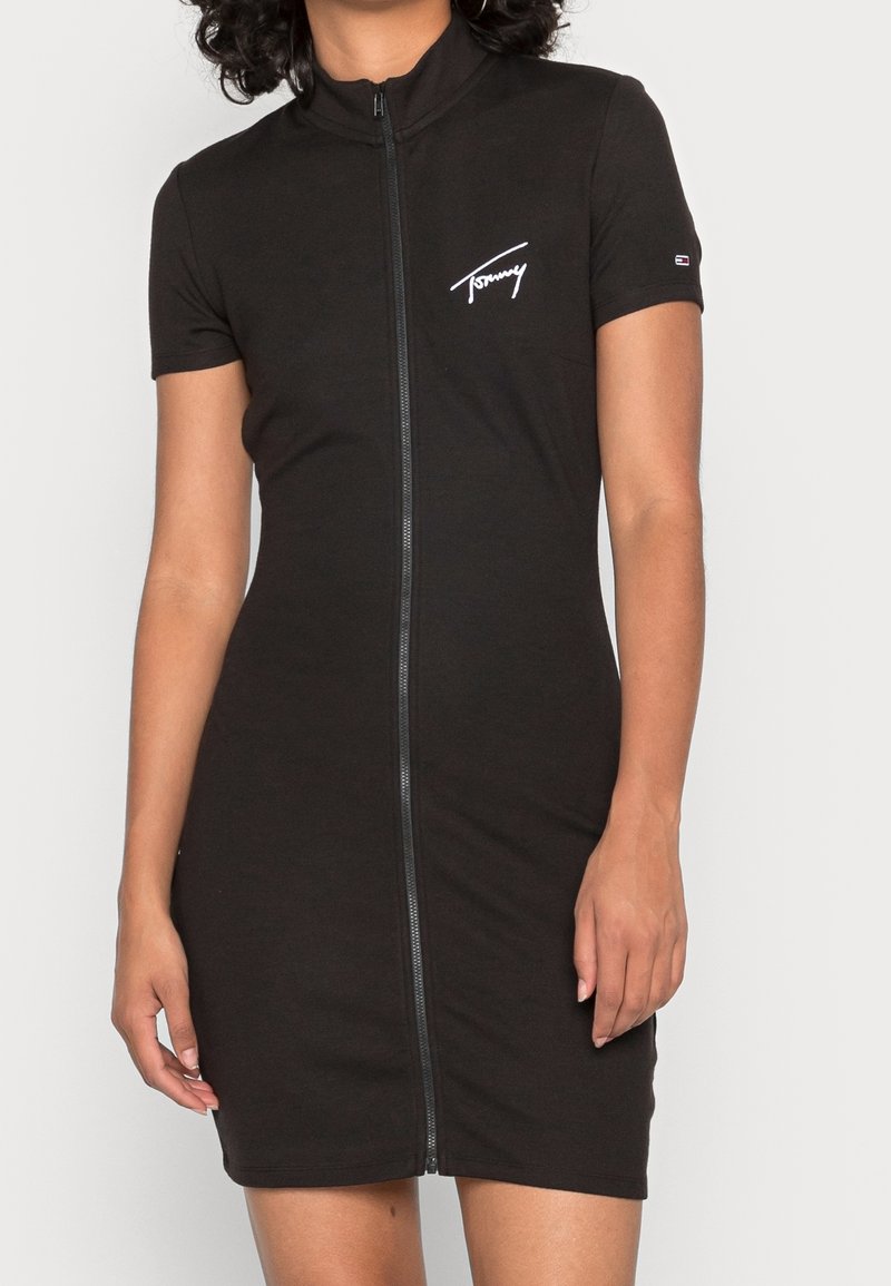 Robe courte noire ajustée à manches courtes avec fermeture éclair à l'avant et logo blanc "Tommy" sur la poitrine, portée par une personne aux cheveux bouclés.
