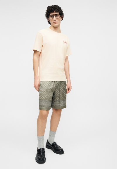 Νεαρός άντρας με σγουρά μαλλιά και γυαλιά, φοράει ανοιχτό ροζ μπλουζάκι, βερμούδα χακί με σχέδια, γκρι κάλτσες και μαύρα μοκασίνια, όρθιος.