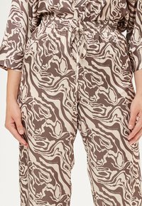 Bruine en crèmekleurige loszittende broek met een elastische tailleband en trekkoord. Voorzien van een abstract patroon en zijzakken.