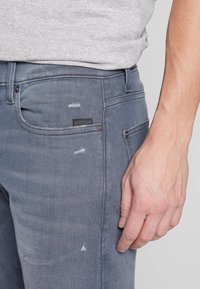 Grå denimjeans med slim fit, slitna detaljer och en svart logotyppatch på bakfickan. Texturen verkar slät och något använd.