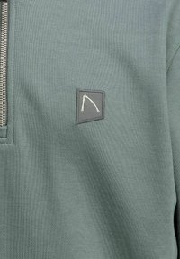 Pullover in tessuto verde opaco e texturizzato con design a mezza zip; presenta una toppa rettangolare con un logo bianco a linea.