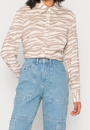 Kobieta w beżowo-białej koszuli na guziki w zwierzęcy wzór zebry, wpuszczonej w jasnoniebieskie, wysokowarstwowe spodnie typu cargo z kieszeniami.