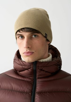 Beanie beige lavorato a maglia con una texture a coste, indossato sopra un giubbotto imbottito marrone con colletto alto e dettagli in zip nera.