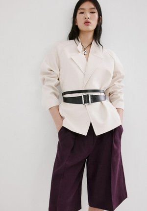 Vrouw draagt een oversized crèmekleurige blazer, getailleerd met een brede zwarte riem, en losse donkerpaarse culottes, staand met handen in de zakken.