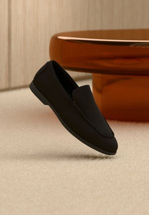 Mocassin noir sans lacets flottant au-dessus d'un tapis clair avec une table moderne incurvée de couleur ambre brun en arrière-plan.
