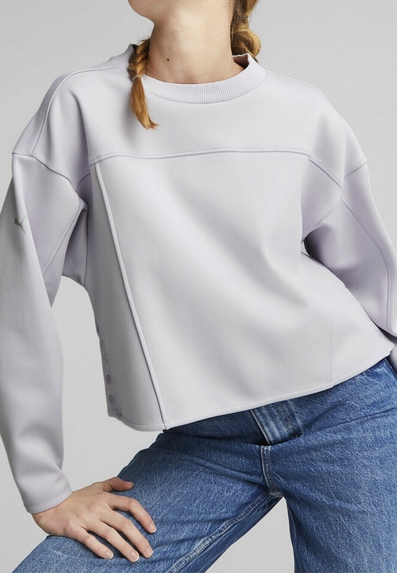 Felpa corta lilla con maniche lunghe, dettagli cuciti e bottoni a pressione all'orlo. Abbinata a jeans in denim blu.