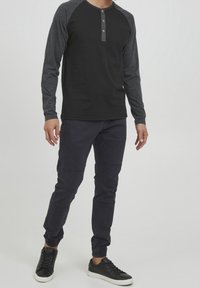 Sort langærmet henley-trøje med grå raglan-ærmer og fem knapper ved halsudskæringen. Kombineret med mørkeblå bukser og sorte sneakers.