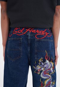 Blue denim jeans with red "Ed Hardy" embroidered waistband, featuring a colorful dragon design with flames on the right back pocket.